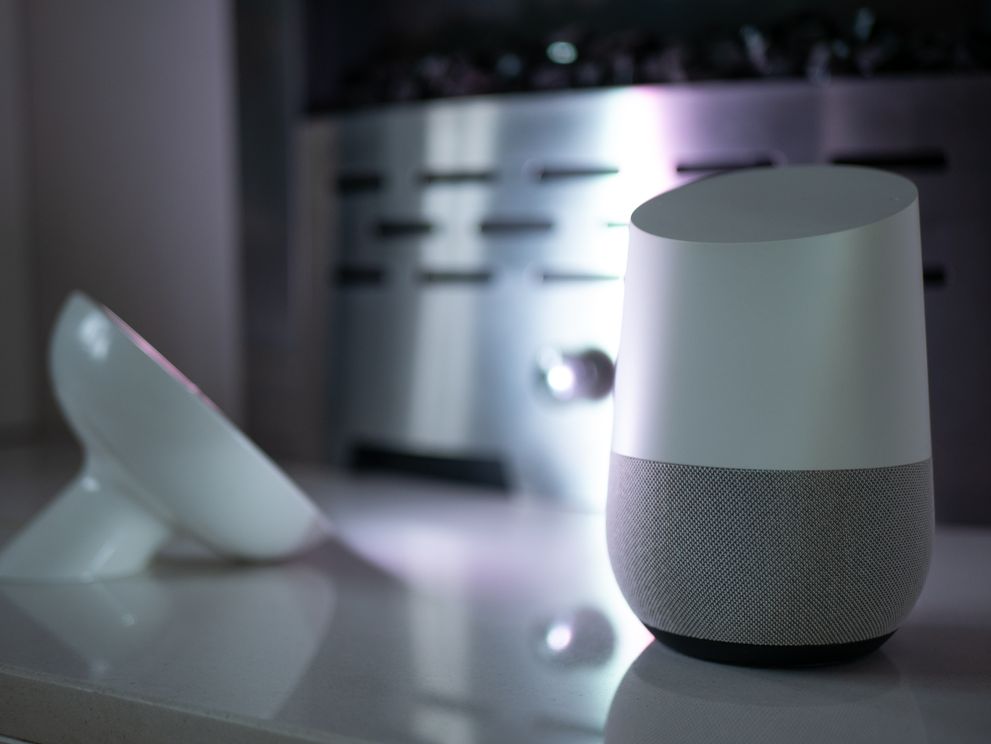 Google Home Sprachassistent und Philips Hue Bloom Tischleuchte im Vordergrund einer bunt beleuchteten Wohneinrichtung